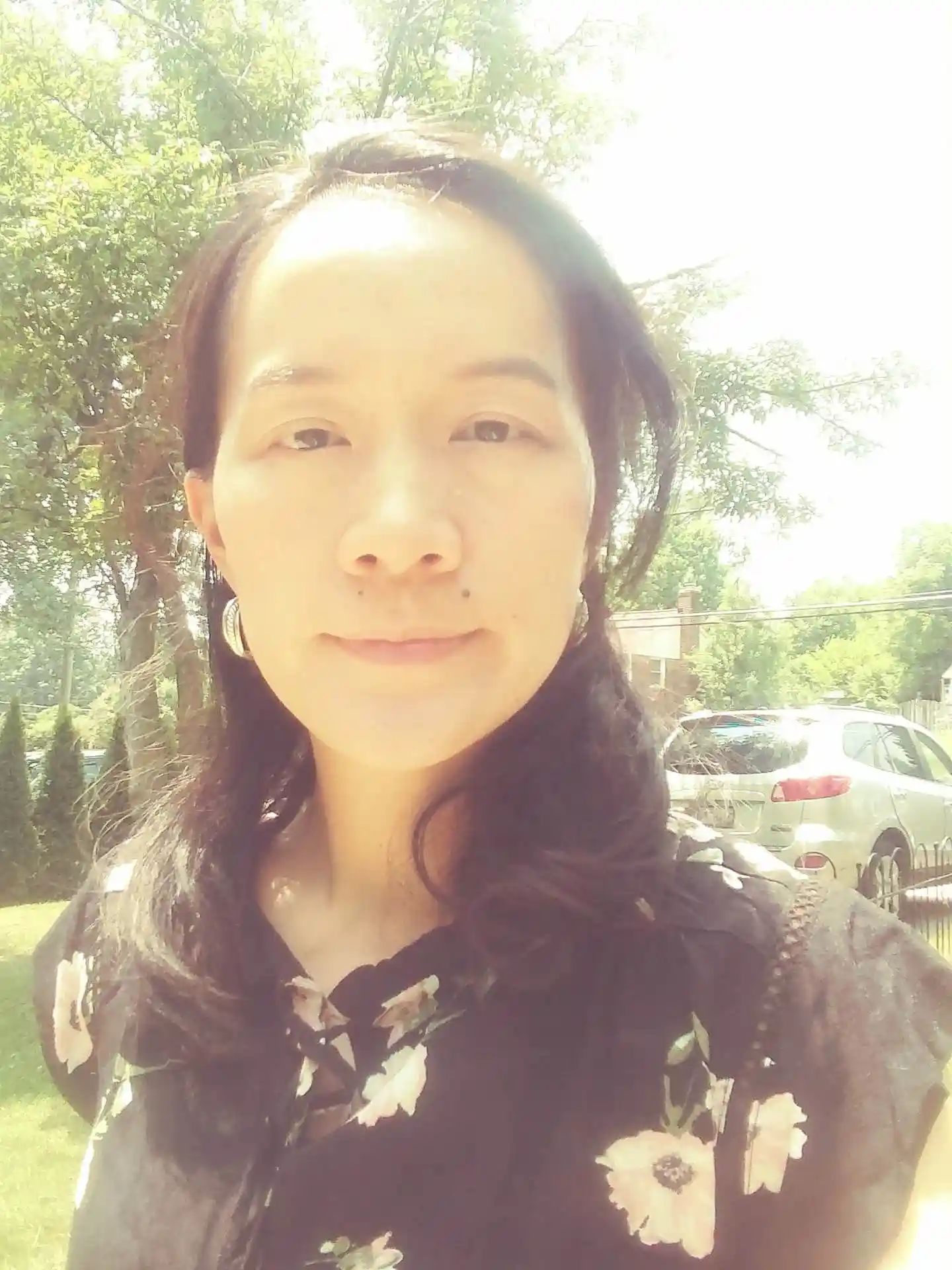 Portrait of a woman with long dark hair, wearing a floral top, standing outdoors with trees and a car in the background.