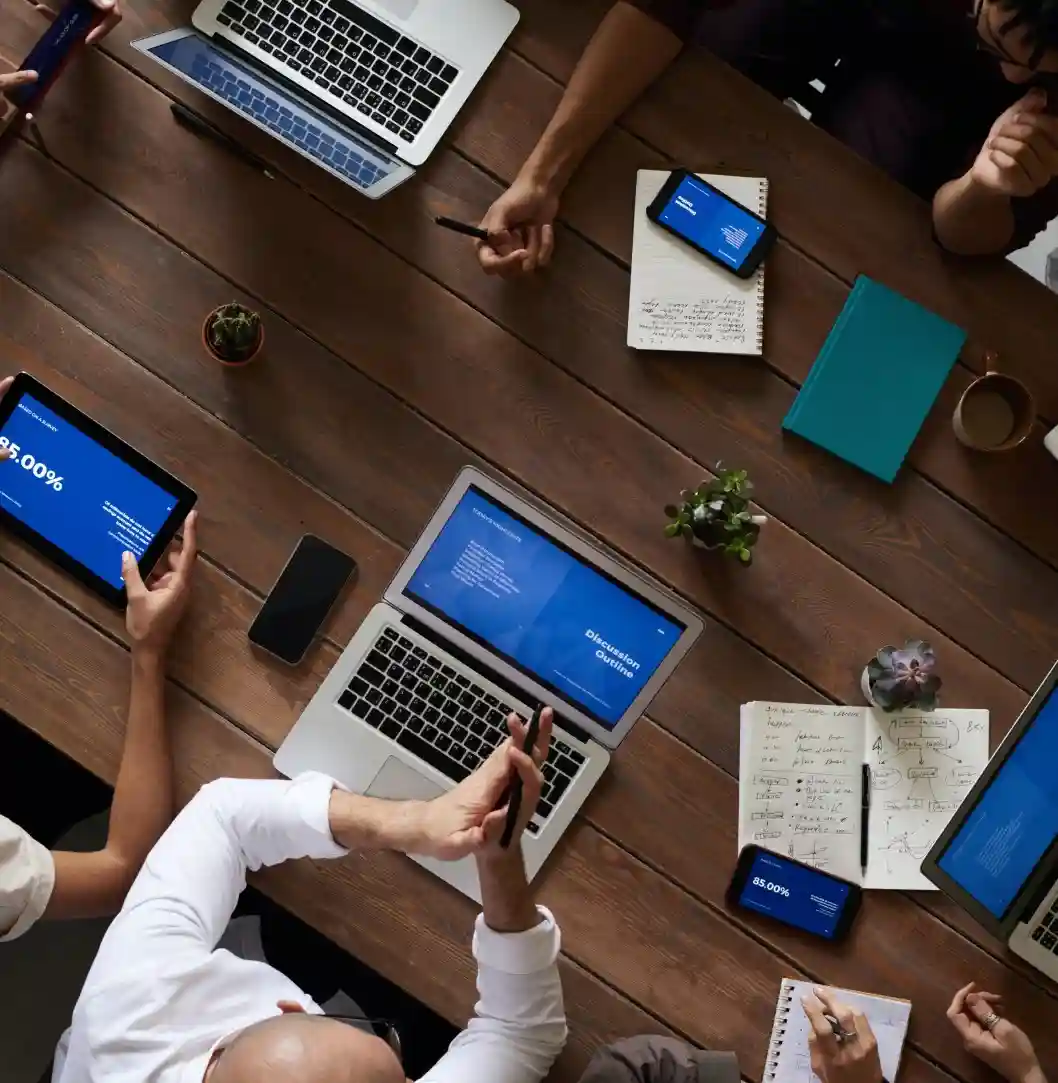 A group of people in a meeting, working together with laptops, tablets, and phones on a wooden table, with a notebook open showing handwritten notes and diagrams.