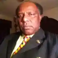 A man dressed in a dark suit with a colorful tie, standing with a serious expression in a room with a dark background.