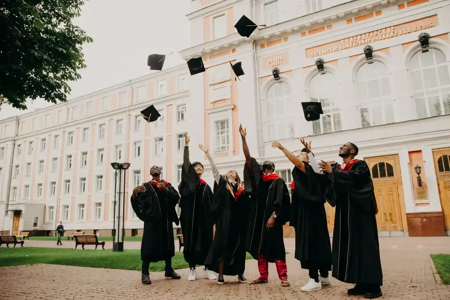 A group of diverse graduates in black gowns throwing their mortarboards into the air in front of a grand academic building.
