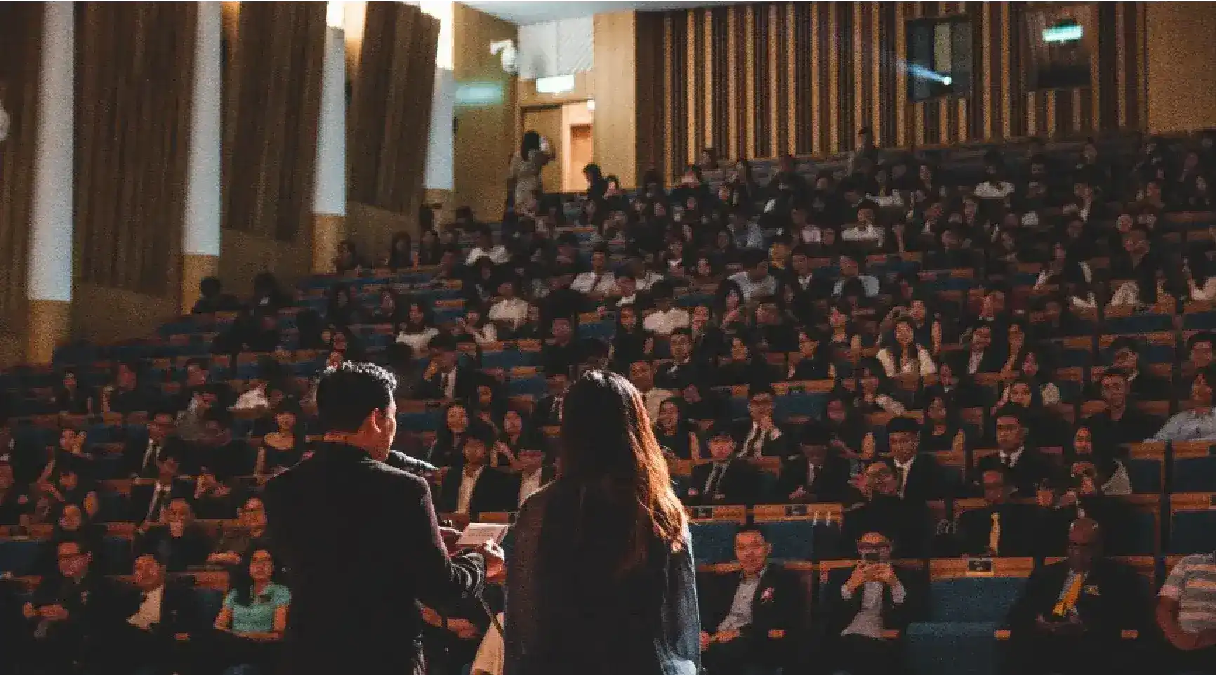 A view from behind two speakers on a stage looking out at a large, seated audience in a tiered auditorium.