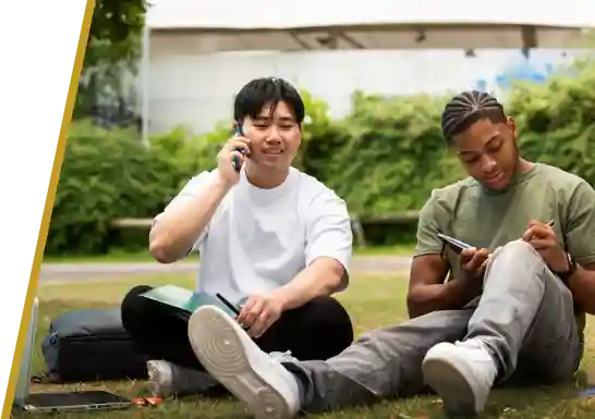 Two students sitting on the grass in a park; one is talking on the phone, while the other is writing on a notepad.
