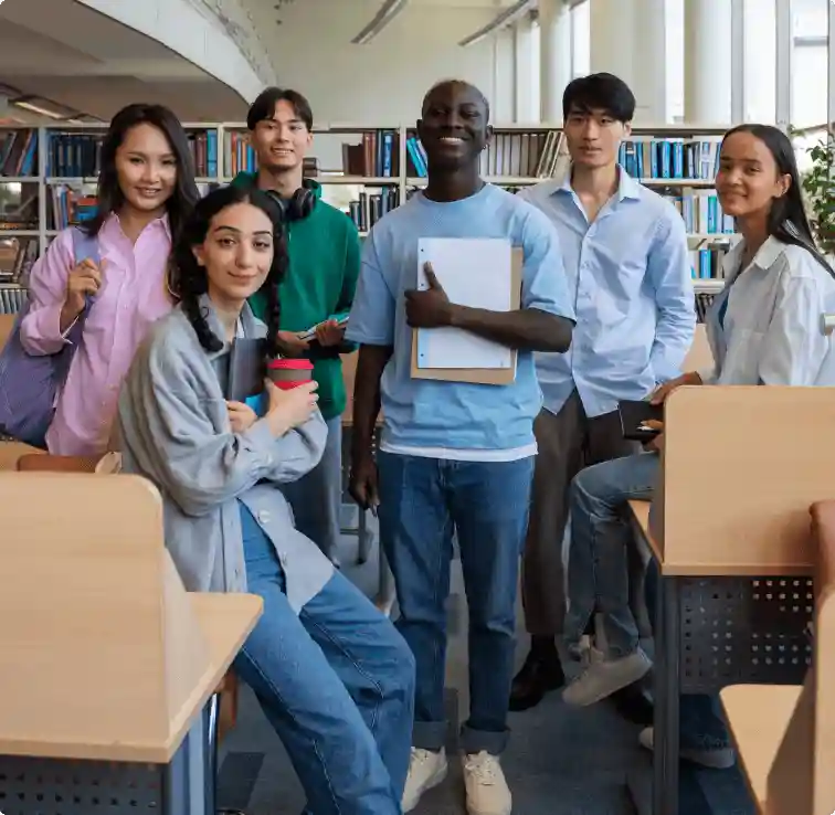 A diverse group of six students standing together in a library, smiling and holding various books and notebooks.