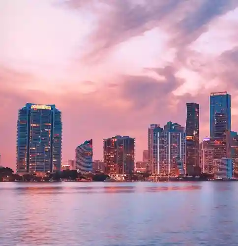 A city skyline at sunset with tall buildings and colorful sky reflected on the water.