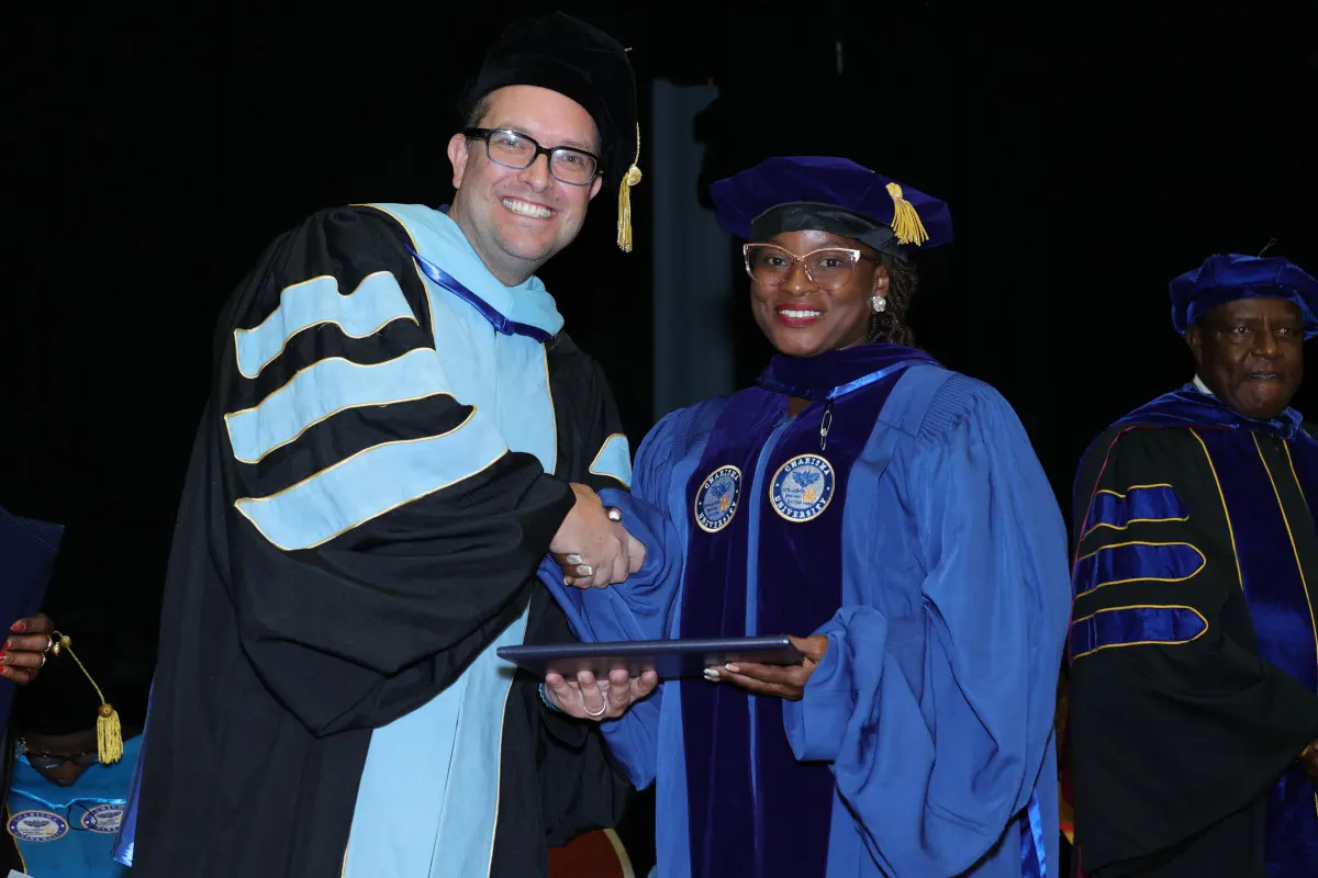 The image shows two individuals shaking hands during a graduation ceremony, both wearing academic regalia.